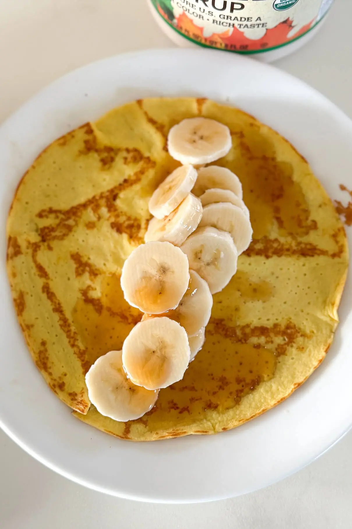 A high-protein pancake on a plate, topped with a line of banana slices and drizzled with golden maple syrup, capturing a close-up of the delicious details.